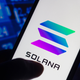 A silhouette holds the Solana logo on a smartphone amid a backdrop of stock numbers.
